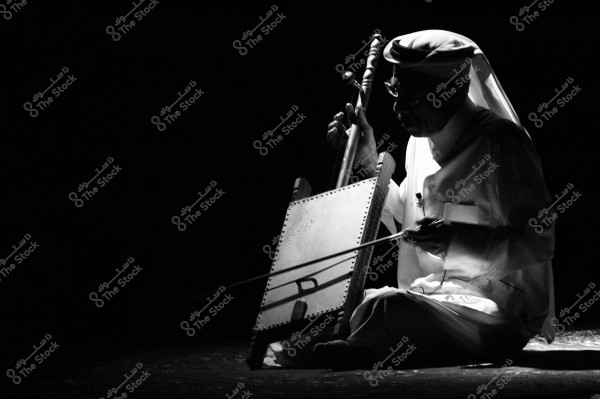 A black and white photo of a man sitting in darkness, playing a traditional musical instrument. The man is wearing an Arabic thobe and a head cover, and the instrument appears to be a rababah. The lighting focuses on the man and his instrument, creating a dramatic effect.