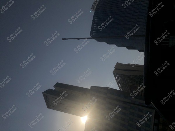 صورة لمشهد حضري يظهر فيه انعكاس ضوء الشمس خلف مجموعة من المباني العالية. البنايات مختلفة التصميم, وبعضها يبدو مغطى بالزجاج, مما يعكس الضوء بشكل جميل على السماء الزرقاء الصافية.