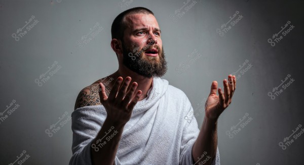 A bearded man wearing a white robe appears in a contemplative or prayerful pose. There is a large tattoo on his arm, and his head is tilted slightly upward. The background is a simple gray.