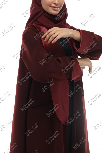 Image of a woman wearing a long burgundy abaya with a matching hijab. The abaya appears elegant with wide sleeves. The image focuses on the upper and middle part of the woman\'s body without showing her full face. The background is simple white.