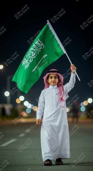 A young boy wearing a white thobe and a red and white checkered headscarf, holding the flag of Saudi Arabia. He is standing on a lit street at night with circular bokeh lights in the background.