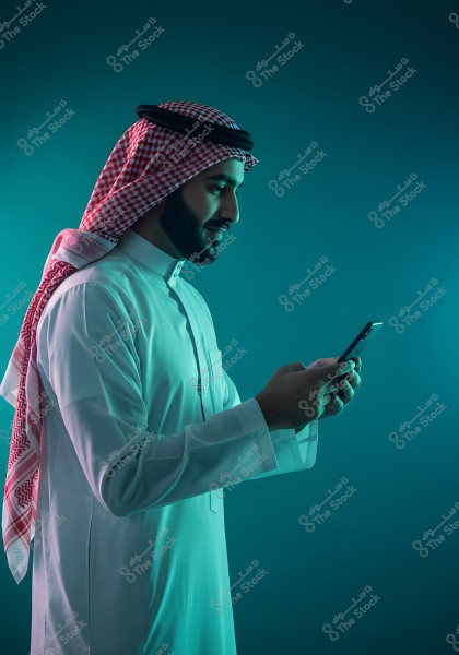 A portrait of a man wearing traditional Saudi attire, including a white thobe and a red ghutra with a black agal, looking at his mobile phone against a calm blue background.
