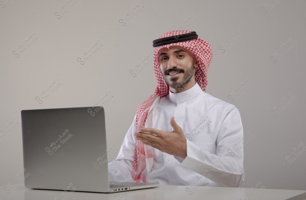 An image of a man sitting in front of a laptop on a table, wearing a traditional white thobe and a red keffiyeh with a black agal. He is smiling and gesturing with his hand. The attire suggests he is from the Gulf region.