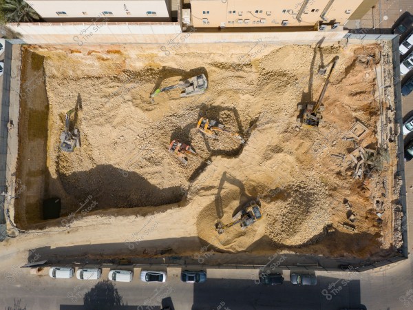 Aerial view of a construction site featuring a large excavation pit surrounded by concrete walls. Five excavators are visible in the pit, moving soil. Parked cars line the street surrounding the site. Residential buildings are visible in the background.