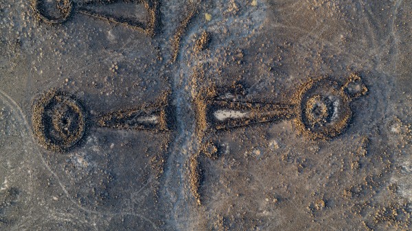 Aerial view of a large stone formation on the ground, featuring three large circles connected by straight lines, with scattered stones and desert terrain surrounding it. The colors present an overall earthy and brown appearance.