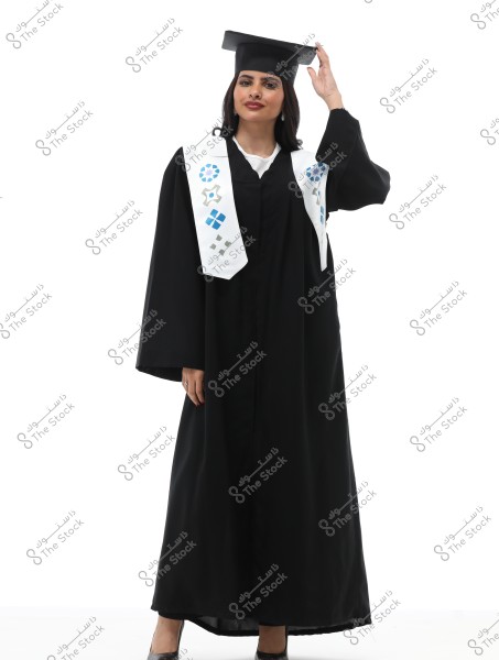 An image of a woman wearing a black graduation gown with a decorated white stole. She is wearing a graduation cap and has her hand on the cap. She stands confidently and smiles at the camera against a white background.