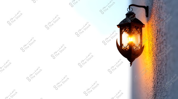 A traditional metal lantern with intricate decorative designs, glowing brightly with an orange hue that reflects off the adjacent gray wall. The lantern is mounted on a metal wall bracket, and the background of the image features a stark white empty space, enhancing the contrast of the warm light emitted from the lantern.