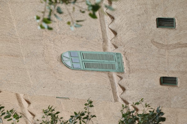 The image shows a decorated mud wall facade with a traditional green wooden window featuring glass sections at the top. The wall is adorned with designs inspired by traditional architecture, with additional small vent windows nearby. Some foliage is visible in the foreground.