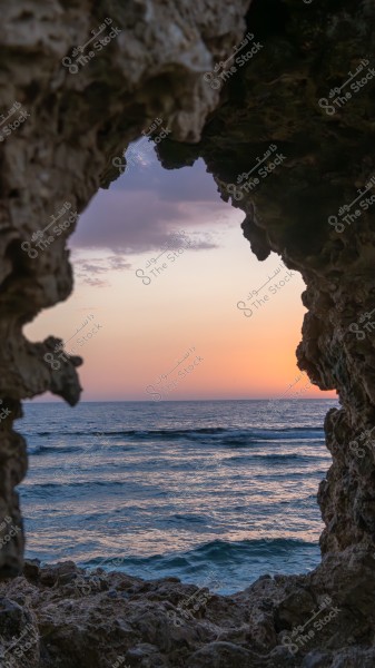 A scenic view of the sea at sunset, seen through an opening in a rocky cave. The horizon is colored with shades of orange and purple as waves gently lap the shore. The rock formations frame the scene dramatically.