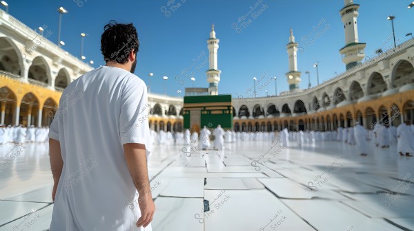 The image shows a man wearing a white garment standing in the courtyard of the Great Mosque in Mecca, with the Kaaba in the background. The courtyard is surrounded by architectural arches and two minarets rising into the clear blue sky. A large number of people, also in white attire, are present.