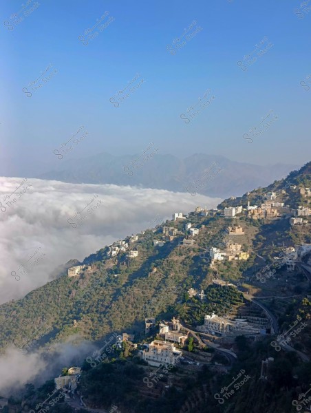 صورة لقرية جبلية تقع على منحدر مغطى بالخضرة والمباني. تنتشر المنازل البيضاء فوق التلال الخضراء, مع وجود سحب تغطي أسفل التلال وتبدو كبحر من الضباب. السماء الزرقاء الصافية تمتد في الخلفية, مما يبرز جمال الطبيعة الريفية.