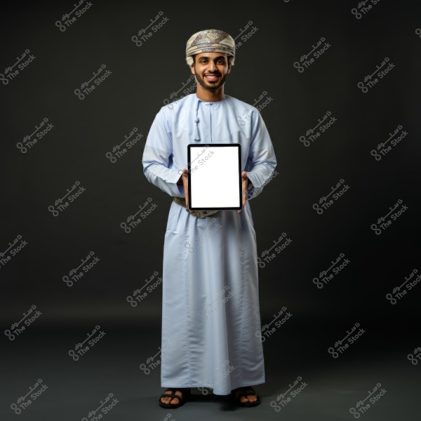 A portrait of a man wearing traditional Omani attire, including a white dishdasha and a decorative Omani turban. The man is standing and smiling, holding a tablet with a blank screen. The background is dark, highlighting the details of the clothing and the device.