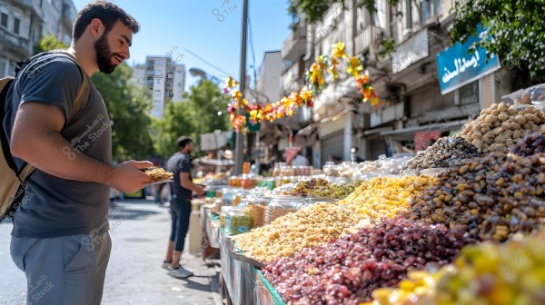 The image shows a man standing in an outdoor market where a variety of nuts and dried fruits are displayed on a table. The man is wearing a gray shirt and has a beige backpack over his shoulder, holding a plate with some goods in his hand. In the background, there are signs, shops, and other people walking around the market. Colorful decorations hanging above the stalls add a festive atmosphere to the setting.