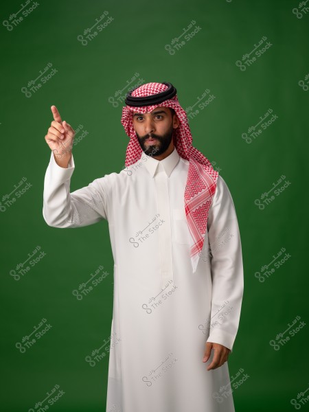 A portrait of a man wearing traditional Gulf attire, including a white thobe and a red and white checkered scarf (shemagh) with a black agal. He stands against a green background, raising his right index finger, appearing focused.