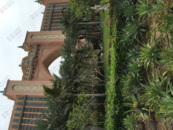 The image shows a large building with luxurious architectural design resembling traditional Arabic style, featuring a large arch and ornate roofs. In the foreground, there is a garden filled with green plants and palm trees, with carefully arranged grassy areas.