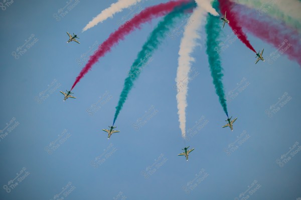 An image showing a formation of six aerobatic planes flying in the blue sky, creating smoke trails in white, red, and green colors. The planes are in an impressive and harmonious coordination, forming a spectacular air show.