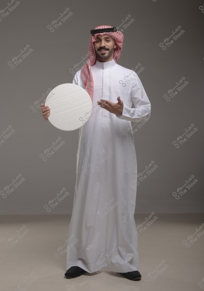 Portrait of a man standing in a studio, wearing a white thobe and a red and white checkered ghutra with a black agal, indicative of traditional Saudi clothing. The man is holding a white circular object with his right hand and gesturing towards it with his left hand while smiling.