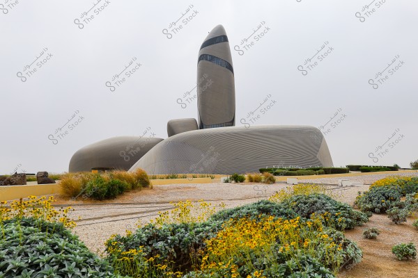 صورة لمبنى مركز الملك عبد العزيز الثقافي العالمي, المعروف باسم \"إثراء\", في الظهران, المملكة العربية السعودية. يتميز المبنى بتصميمه المعماري العصري والفريد من نوعه, حيث يتكون من عدة هياكل منحنية وملساء بلون رمادي. يحيط المبنى حديقة تحتوي على أزهار ونباتات خضراء, مما يضفي لمسة من الطبيعة على المنظر العام.