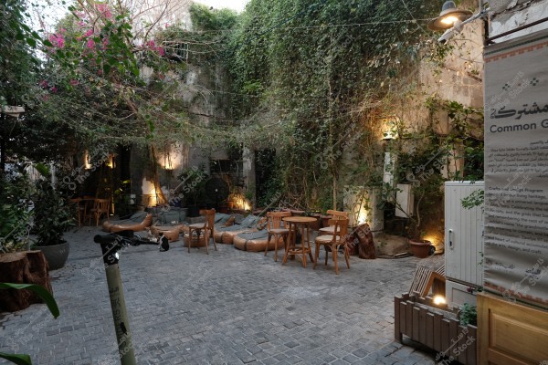 فناء خارجي هادئ يحتوي على طاولات وكراسي خشبية محاطة بالنباتات الخضراء الزاحفة، مع إضاءة دافئة ولوحة مكتوب عليها \"المشتركة\" باللغتين العربية والإنجليزية.