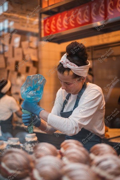 A baker wearing blue gloves and a white headband uses a piping bag to decorate pastries in a bakery.