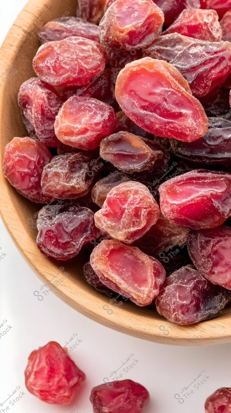 A wooden bowl filled with an assortment of dried dates in pink and dark brown colors. The dates are shown with clear details and a sugary texture, with a few pieces scattered on a white surface next to the bowl.