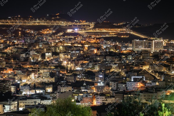 An image of a city at night showcasing an urban scene lit by the bright lights of buildings and streets. In the background, dark hills and illuminated roads create a beautiful contrast between the lit areas and shadows. The buildings appear densely packed, indicating a high population density.