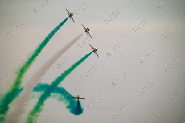 The image shows four fighter jets flying in an aerial formation with colored smoke trailing behind them. The smoke is green and white, creating a visually striking display against the sky backdrop. The jets are flying in diverging directions with intersecting smoke trails.