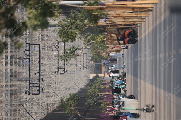 An image of a modern street featuring a neatly constructed pathway lined with neatly arranged electric scooters. Small trees are evenly planted along both sides of the path, with a few individuals in traditional clothing in the background. Arabic writing is visible on the walls behind the people. The atmosphere appears sunny and calm.