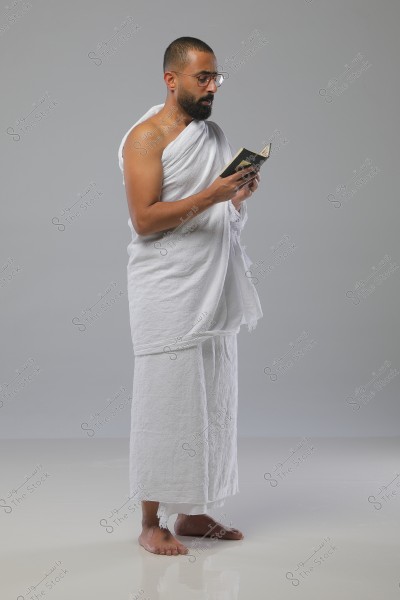 A portrait of a man standing against a gray background, wearing the traditional Ihram attire for Hajj, consisting of two pieces of unstitched white cloth. He is barefoot, wearing glasses, and reading from a small book in his hands. His expression appears focused and meditative.