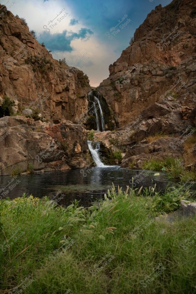 An image of a waterfall cascading between rugged rocky mountains, with water flowing into a small pool surrounded by green plants and grass. The background shows a cloudy sky, adding to the natural beauty of the picturesque scene.