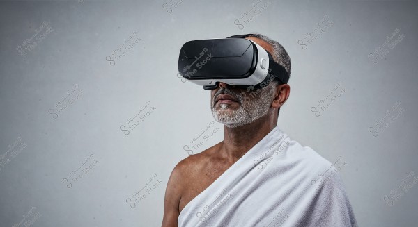 Image of a man wearing virtual reality goggles. The man is standing against a gray background, dressed in a piece of white cloth draped around his body similar to Ihram attire.
