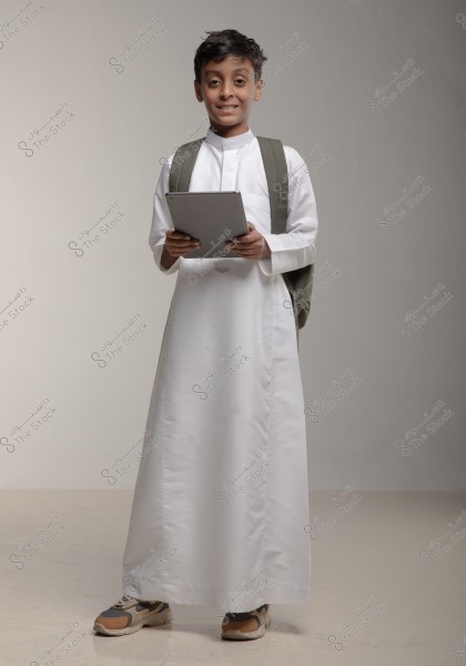 A portrait of a smiling boy dressed in a traditional white thobe, suggesting a likely origin from Saudi Arabia or the Gulf region. He is holding a tablet and wearing a green backpack. His shoes are sporty with a modern design. The background is neutral, drawing attention to the child.