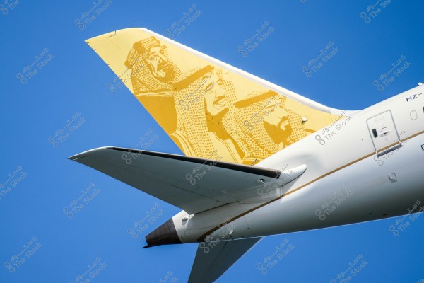 The image shows the tail of an airplane featuring artwork of three men wearing traditional Saudi attire, including the thobe and shemagh. A clear blue sky forms the background.
