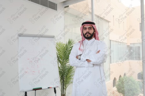 رجل سعودي يرتدي الزي السعودي التقليدي يقف مكتوف اليدين أثناء إجتماع في العمل نهارًا