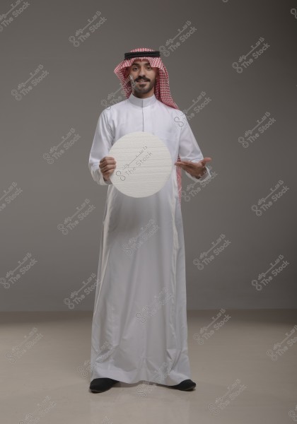 An image of a man wearing a traditional white thobe and a red and white checkered shemagh with an agal. He is holding a blank circular board in front of him and pointing at it with one hand. The background is simple and gray.