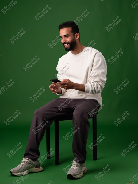 A man sitting on a chair against a green background. He is wearing a white sweatshirt, dark pants, and gray sneakers. He is holding a mobile phone and smiling naturally.