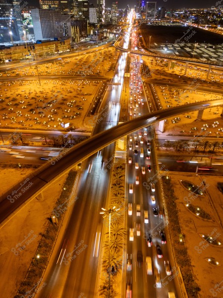 An aerial night view of a city illuminated with bright lights. The image shows a highway intersected by bridges, crowded with cars creating light trails due to a long exposure. Tall buildings surround the road, with the city lights sparkling in the background.