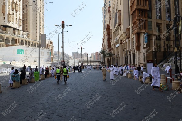 The image shows a busy street in an urban area, with people wearing white ihram garments, suggesting they may be in Saudi Arabia near the Haram area. Tall buildings line both sides of the street. Some individuals are wearing yellow vests, possibly workers or volunteers, and the street appears lively with activity.