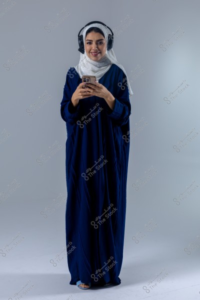 صورة لامرأة ترتدي عباءة زرقاء داكنة وحجاب أبيض, وتضع سماعات رأس سوداء كبيرة الشكل. تقف مبتسمة وتمسك بهاتف بيديها, في خلفية بيضاء.