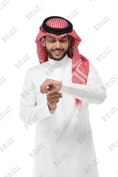 A portrait of a man wearing a white thobe and a red-checkered ghutra with an agal. He appears to be looking at his wristwatch with a calm smile. The attire suggests he is from Saudi Arabia.