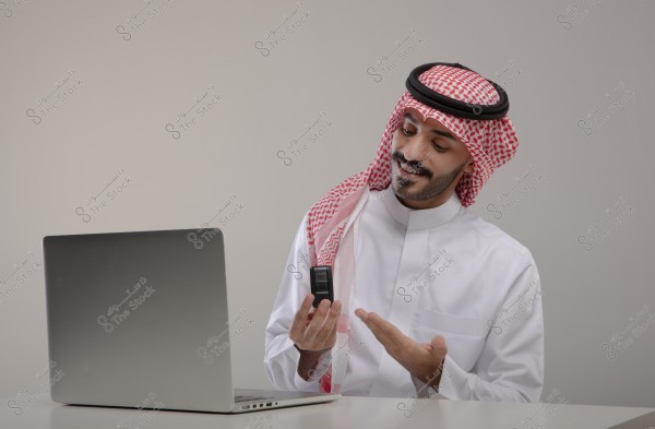 A portrait of a man wearing a Saudi thobe and shemagh, sitting in front of a laptop. He appears to be interacting, holding a remote control in his right hand while gesturing with his left hand. The background is simple, emphasizing the focus on the subject.