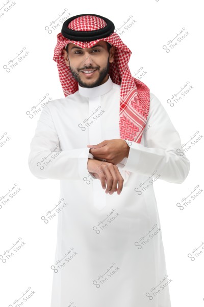 The image shows a man wearing traditional Saudi attire, consisting of a white thobe and a red and white checkered shemagh. The man is smiling and adjusting the sleeves of his thobe. The background is white.