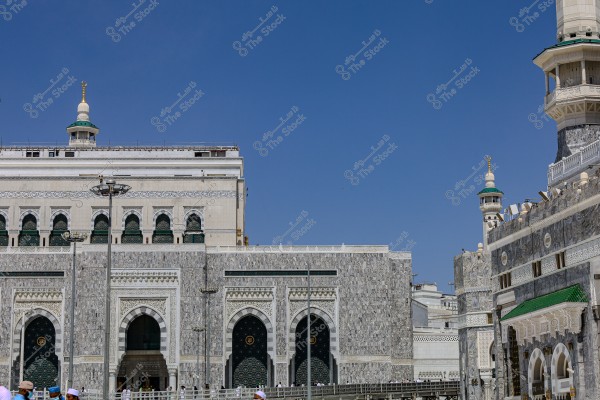 صورة تظهر جزءًا من المسجد الحرام في مكة المكرمة, السعودية. تتضمن الصورة تفاصيل العمارة الإسلامية, مع مآذن مزخرفة وقباب وأبواب كبيرة مزخرفة. السماء زرقاء وصافية, وهناك بعض الأشخاص يظهرون في الزاوية السفلية.