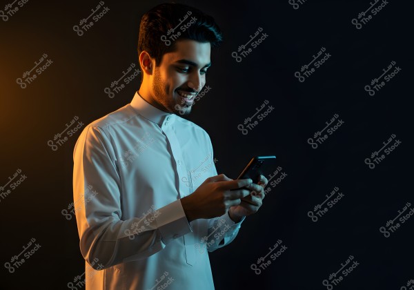 A photo of a man wearing a white thobe browsing a smartphone. The background is dark, highlighting the light on his face. He seems to be enjoying what he sees on the phone.