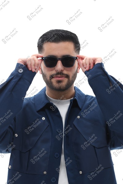 A man wearing sunglasses holding the sides with his hands. He is dressed in a dark blue shirt with metal buttons and stands against a plain white background. The photo appears to be taken in a portrait style.
