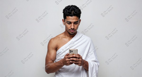 A portrait of a man wearing a white wrap similar to Ihram, standing against a white background, holding a smartphone in his hands. His facial expression is focused as he looks at the phone. The wrap appears to be made of soft, comfortable fabric.