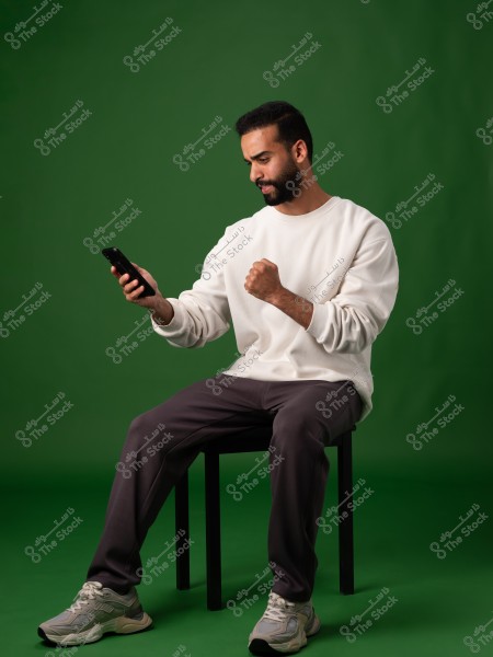 Image of a man sitting on a chair against a green background. He is wearing a white sweater, dark pants, and gray sneakers. He is holding a phone in his left hand and appears happy or triumphant as he clenches his right fist in a gesture of joy.
