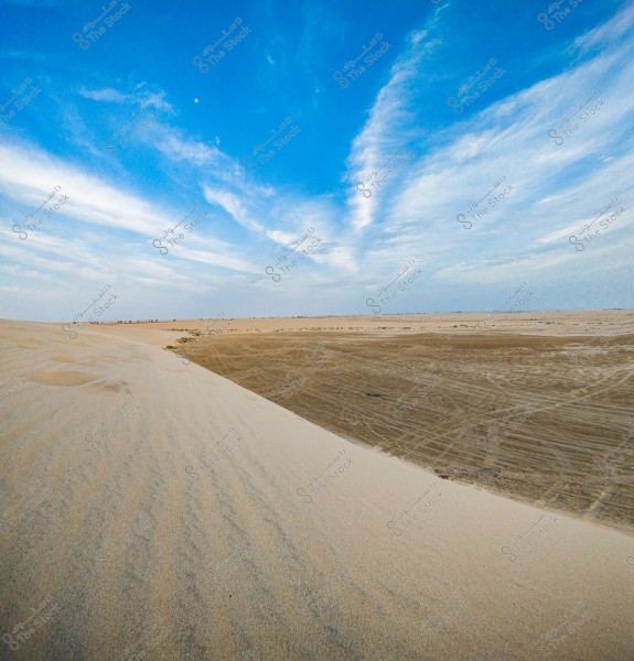 An image showing a desert landscape with a clear blue sky and wispy clouds. The sand stretches toward the horizon, with tire tracks visible on the darker sand.