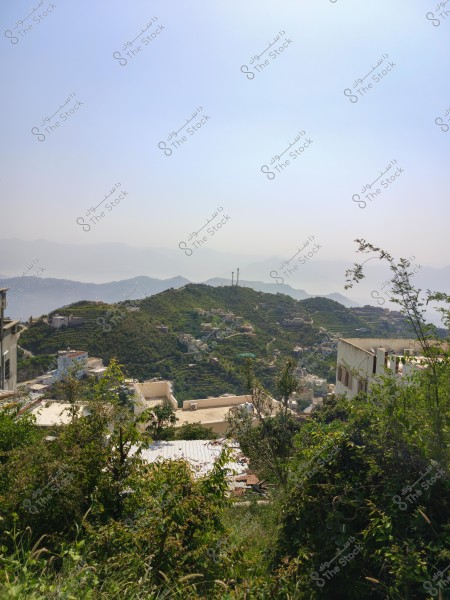 A natural landscape overlooking green hills with scattered buildings on the mountain slopes. There are green trees and plants in the foreground, while misty mountains appear in the background with a clear blue sky.