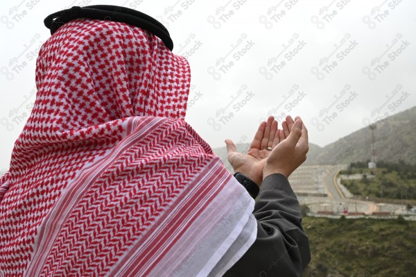 A shot of a Saudi man wearing the Saudi dress, praying and standing on one of the green mountains.